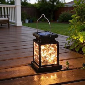 May include: Black solar-powered lantern with a silver handle, featuring a small solar panel on top. The lantern's interior is filled with warm white fairy lights. The lantern is sitting on a wooden deck.