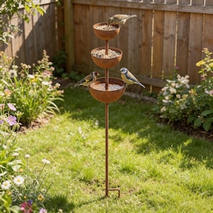 May include: A three-tiered, weathered metal bird feeder with bowls for food and water. Several small birds are perched on the feeder, feeding. The feeder is set in a garden with grass, flowers, and a wooden fence in the background.