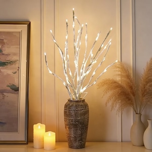 May include: A decorative arrangement featuring white birch branches illuminated with small white lights, displayed in a textured brown vase. Two lit candles and a framed painting are in the background, creating a warm ambiance.