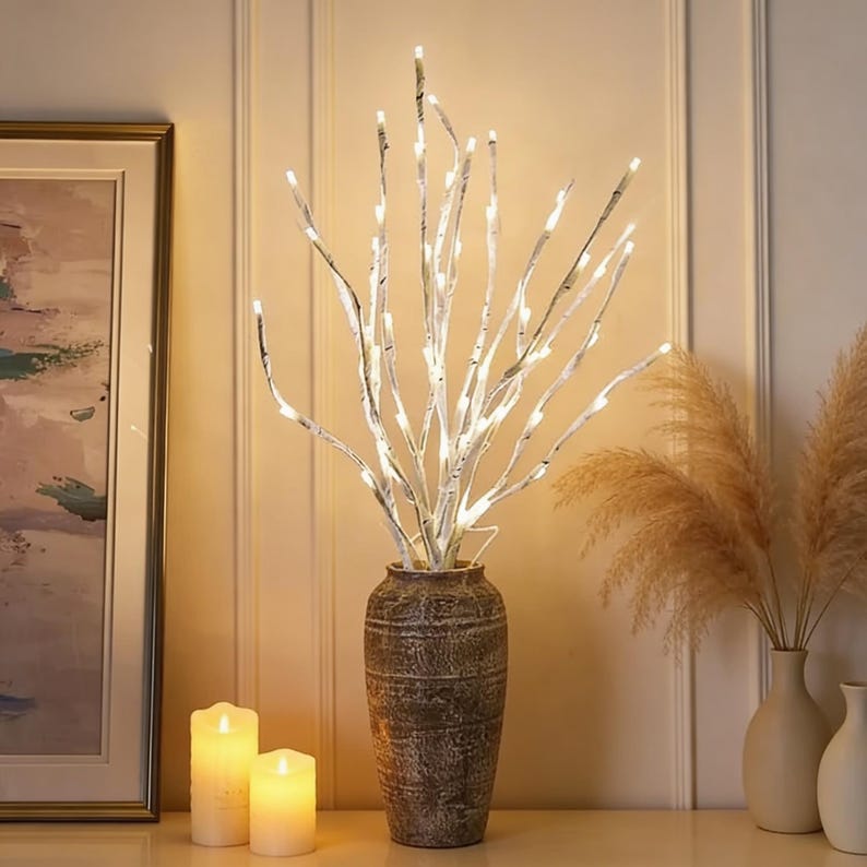 May include: A decorative arrangement featuring white birch branches illuminated with small white lights, displayed in a textured brown vase. Two lit candles and a framed painting are in the background, creating a warm ambiance.