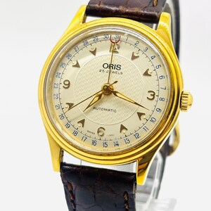 May include: An Oris automatic wristwatch with a gold-tone case and a textured cream dial. The watch features a date pointer hand with a red crescent tip, Arabic numerals at three and nine o'clock, and a dark brown embossed leather strap.