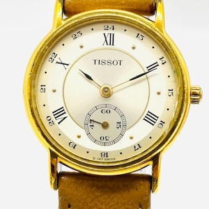 May include: A Tissot wristwatch with a gold-tone round case and a tan leather strap. The cream-colored dial features black Roman numerals at the 12, 10, 8, 6, and 4 o'clock positions, Arabic numerals for 24-hour time, and a small seconds sub-dial.