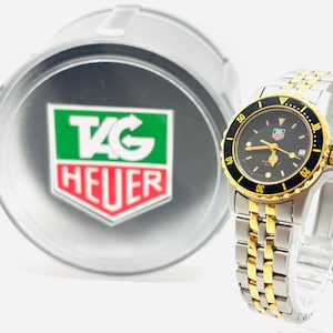 May include: A TAG Heuer watch with a black dial, gold bezel, and a two-tone silver and gold bracelet. The watch is displayed next to a silver container with the TAG Heuer logo.