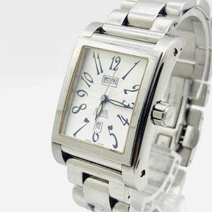 Puede incluir: Un reloj de pulsera plateado con esfera rectangular. La esfera es blanca con números y agujas negras. La correa es de metal. La esfera muestra el día de la semana y la fecha. El reloj está etiquetado como "ORIS AUTOMATIC SWISS MADE".