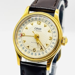 Puede incluir: Un reloj de pulsera automático Oris dorado con esfera crema y correa de cuero marrón. La esfera muestra el logotipo de Oris, 17 rubíes y un indicador de fecha. El reloj tiene un bisel dorado y una correa de cuero marrón.