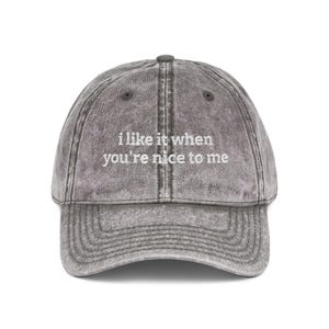 May include: A gray baseball cap with the text "i like it when you're nice to me" embroidered in white. The cap has a curved brim and a button on top. The hat is made of a soft, worn material.