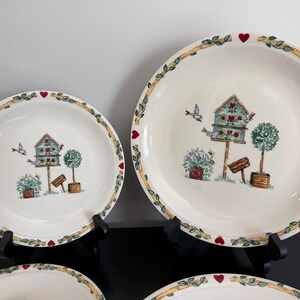 May include: Two round ceramic plates of different sizes featuring a cream-colored base with a painted design of a birdhouse, green topiary trees, and small birds. The rim is decorated with a yellow vine pattern and small red heart accents.