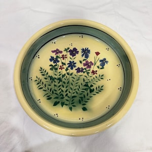May include: Round ceramic serving bowl with a floral design. The bowl features a cream-colored interior with a hand-painted bouquet of blue, purple, and red flowers. The rim is bordered by a dark green band and a light yellow edge.