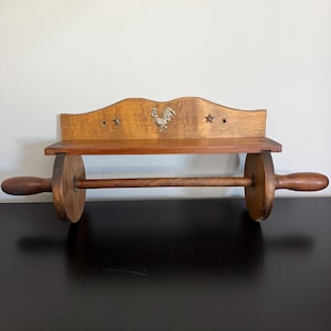 May include: A wall-mounted wooden paper towel holder featuring a shelf with a rooster emblem and two star cutouts. The design incorporates a horizontal rod supported by two circular wooden wheels, finished in a medium brown wood tone.