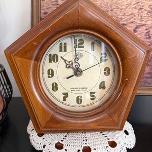 May include: A pentagonal wooden clock with a light-colored face featuring black Arabic numerals and ornate black hands. The clock face displays the text WOODEN FASHION WOOD LAND CO. LTD. and a small logo. The frame is made of polished brown wood.