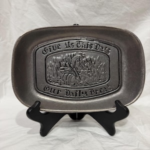 May include: A rectangular, pewter-colored bread plate with the words "Give Us This Day Our Daily Bread" embossed on the front. The plate features a relief image of a person harvesting wheat. The plate is displayed on a black stand.