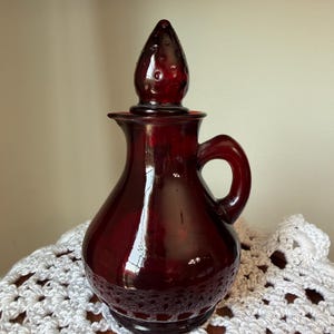 May include: A deep red glass cruet with a rounded body, a small side handle, and a matching pointed stopper. The glass features a smooth, translucent finish with a bulbous base and a narrow neck, reflecting light across its dark crimson surface.