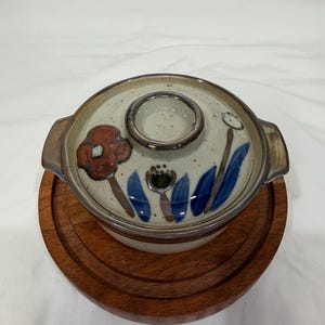 Może przedstawiać: Ceramiczna miska do serwowania z pokrywką i kwiatowym wzorem w kolorze czerwonym, niebieskim i zielonym. Naczynie stoi na okrągłym drewnianym podkładzie. Pokrywka ma okrągły uchwyt.