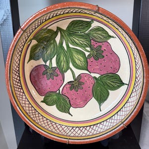 May include: A round ceramic plate featuring a hand-painted design of three pink strawberries with green leaves. The plate has a cream-colored center, a yellow inner border, and a wide terracotta-colored rim with a cross-hatched pattern.