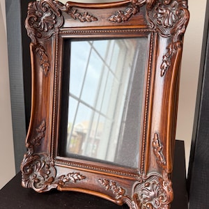 May include: A rectangular picture frame with a dark brown wood grain finish and ornate, raised floral carvings at each corner and along the edges. The frame features a central glass pane and a beaded inner border surrounding the display area.