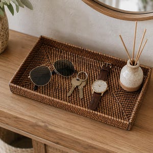 May include: A rectangular woven rattan tray in a dark brown finish containing a pair of aviator sunglasses with dark lenses, a silver metal keyring with two keys, a wristwatch with a brown leather strap, and a small ceramic reed diffuser.