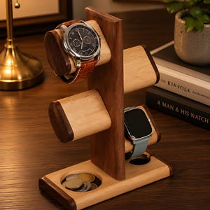 May include: A two-tone wooden watch stand featuring a vertical central pillar with three horizontal cylindrical bars for displaying timepieces. One bar holds a silver analog watch with a brown leather strap, and another holds a smartwatch.