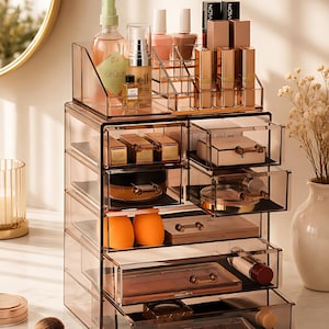 Könnte beinhalten: Ein klarer, mehrstufiger Make-up-Organizer mit mehreren Schubladen und Fächern. Der Organizer ist mit verschiedenen Schönheitsprodukten gefüllt, darunter Lippenstifte, Nagellack und Hautpflegeartikel. Das transparente Design ermöglicht eine einfache Sichtbarkeit des Inhalts.
