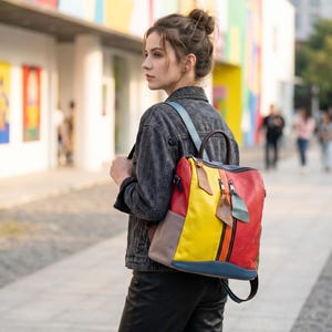 May include: A multi-colored leather backpack with a vibrant design. The backpack features panels of red, yellow, blue, and gray. It has a top handle, adjustable straps, and a front zipper. The bag is worn over the shoulders.