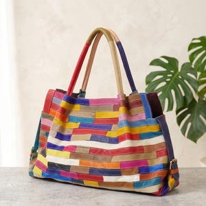 May include: A large, colorful patchwork leather tote bag. The bag features a variety of colors, including red, yellow, blue, and green, arranged in horizontal stripes. It has two top handles and a shoulder strap. The bag is sitting on a gray surface.