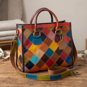 May include: A multi-colored handbag featuring a patchwork diamond pattern in shades of red, blue, yellow, orange, brown, and pink. The bag includes two rounded top handles, a detachable brown fabric shoulder strap, and a blank rectangular leather tag.