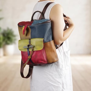 Könnte beinhalten: Ein bunter Lederrucksack mit Patchwork-Design. Die Tasche hat rote, gelbe, blaue und braune Paneele, eine Frontklappe und verstellbare braune Riemen. Sie hat einen oberen Griff und einen Reißverschluss.
