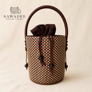 Può includere: Una borsa a cestino cilindrica intrecciata con un motivo a scacchi marrone scuro e crema, un unico manico arcuato e una chiusura a coulisse in tessuto marrone scuro. La borsa è decorata con perline marrone scuro sul cordino e sugli attacchi.