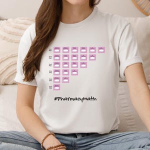 Könnte beinhalten: Ein weißes Kurzarm-T-Shirt mit einem grafischen Aufdruck eines Medikamenten-Zeitplans, bestehend aus rosa Quadraten, die nach Tag und Uhrzeit beschriftet sind, sowie dem Text #PharmacyMath in Schwarz unterhalb des Rasters auf der Vorderseite.