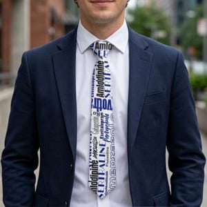 May include: A white necktie featuring a repeating pattern of dark blue and black text, including words like Amlodipine, Sertraline, and Gabapentin, worn with a crisp white button-down shirt and a tailored navy blue blazer jacket.