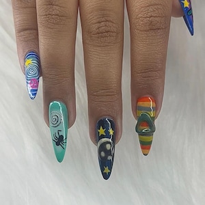 May include: A set of long, pointed fingernails with distinctive nail art. Designs include a swirling galaxy, a spider, a skull with stars, and a striped design with a metal ring. The nails are painted in various colours, including blue, teal, and orange.