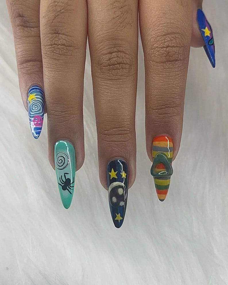 May include: A set of long, pointed fingernails with distinctive nail art. Designs include a swirling galaxy, a spider, a skull with stars, and a striped design with a metal ring. The nails are painted in various colours, including blue, teal, and orange.