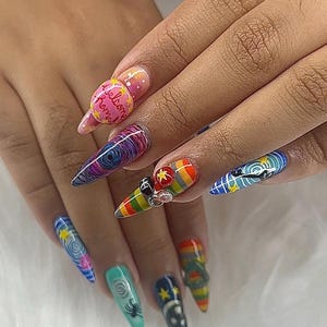 May include: A hand with long, pointed nails, each adorned with intricate nail art. Designs include swirls, rainbows, stars, and a spider. One nail features the text "Welcome Home". The nails are decorated with 3D embellishments.