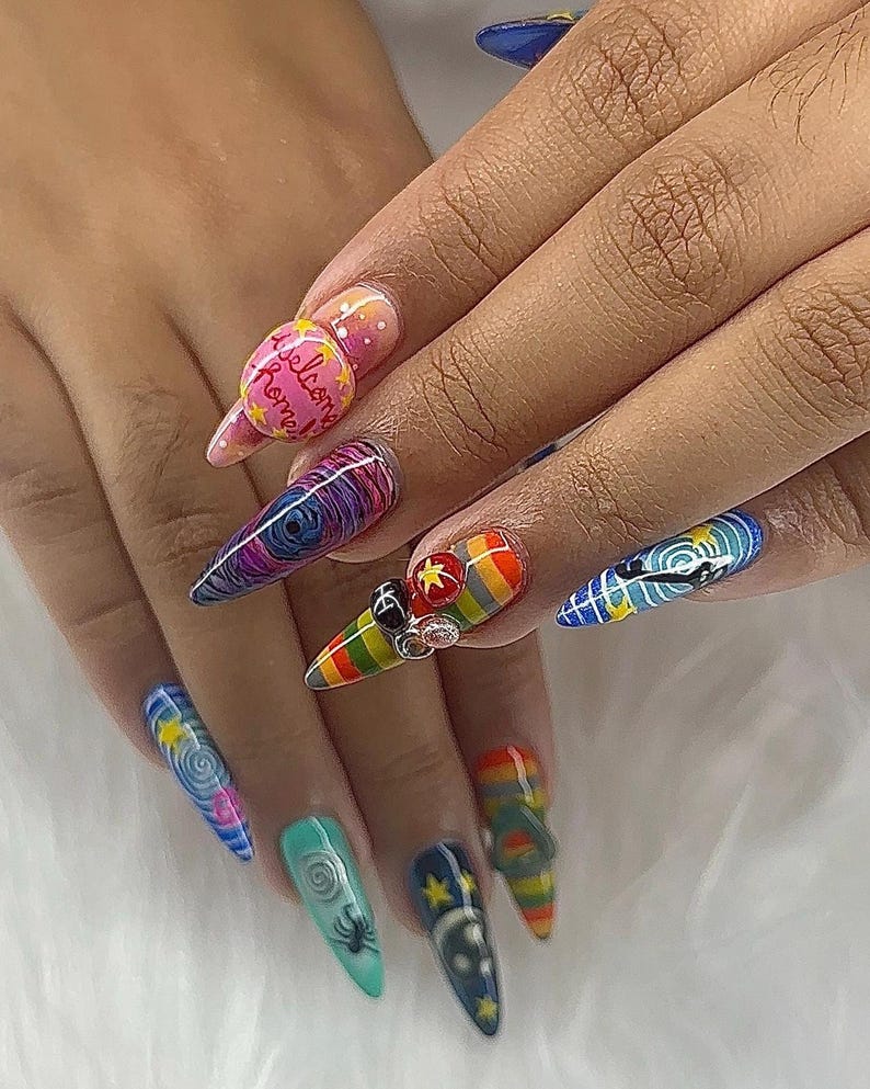 May include: A hand with long, pointed nails, each adorned with intricate nail art. Designs include swirls, rainbows, stars, and a spider. One nail features the text "Welcome Home". The nails are decorated with 3D embellishments.
