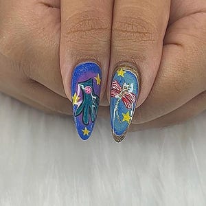 May include: Close-up of two long, oval fingernails with detailed nail art. One nail features a teal creature with yellow stars on a blue background. The other nail has a red and white bow with yellow stars on a blue background, outlined in gold.