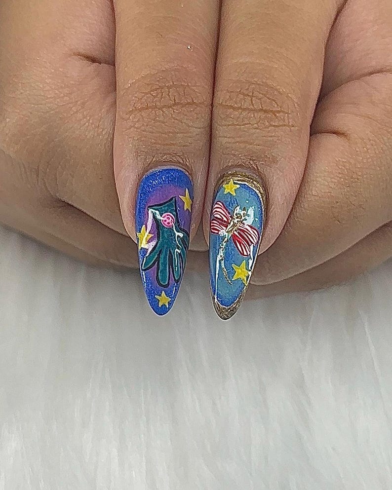 May include: Close-up of two long, oval fingernails with detailed nail art. One nail features a teal creature with yellow stars on a blue background. The other nail has a red and white bow with yellow stars on a blue background, outlined in gold.