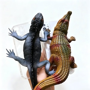 Peut inclure: Une coque de smartphone transparente avec deux figurines de reptiles en plastique fixées dessus. À gauche, un iguane noir texturé aux yeux orange, et à droite, un crocodile texturé marron et vert avec des accents rouges sur le dos et la queue.