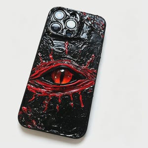 May include: A black smartphone case featuring a textured, three-dimensional surface with a prominent, realistic red eye at the center. The eye is surrounded by raised, glossy red paint strokes that mimic veins and skin folds against a black background.