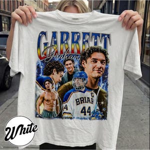 May include: White t-shirt featuring a graphic of Garrett Graham with multiple images of him, including one in a hockey uniform. The design includes the name "Garrett Graham" in a stylized font with a blue, yellow, and red gradient.