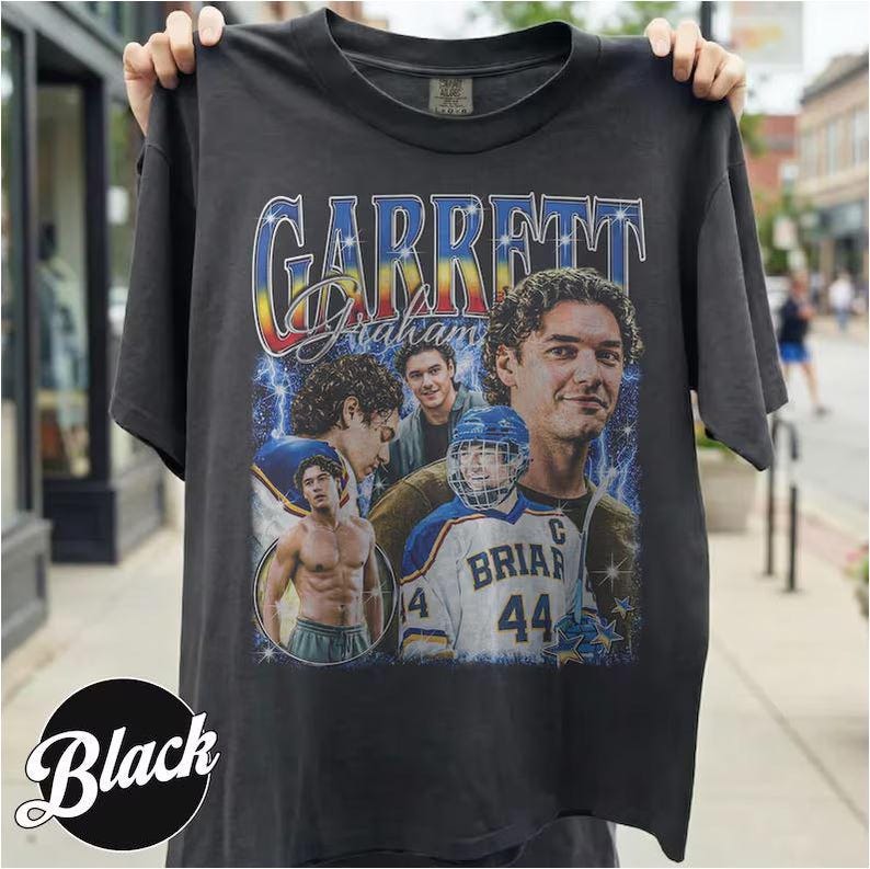 May include: Black t-shirt featuring a graphic design with the name "Garrett Graham" in a retro style. The design includes images of a man in a hockey uniform and other portraits. The shirt is held up by a person.