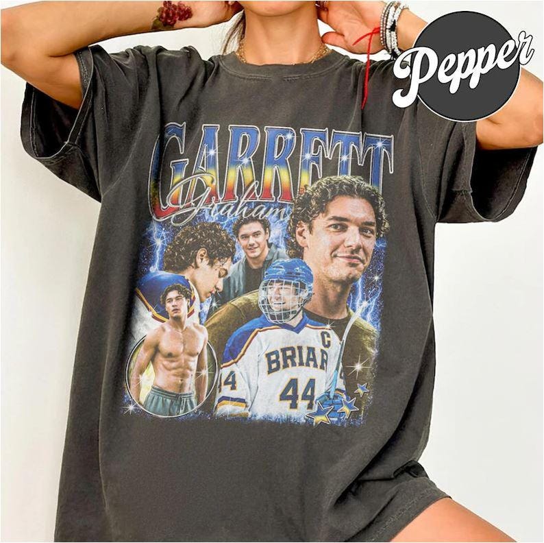 May include: Dark gray t-shirt featuring a collage of images and text. The text reads "Garrett Graham" in a stylized font. The collage includes images of a person in a hockey uniform and other portraits.
