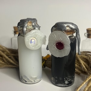 May include: Two glass potion bottles with cork stoppers and silver wax seals. One bottle is filled with a white liquid and the other with a black liquid. Each bottle has a decorative white eye-shaped embellishment.