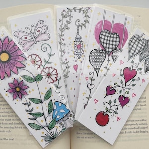 Könnte beinhalten: Ein Set aus fünf rechteckigen Lesezeichen aus Papier mit handgezeichneten Illustrationen. Die Motive umfassen rosa Blumen, einen blauen Pilz, einen Schmetterling, eine hängende Laterne und verschiedene Herzen mit schwarz-weißen Mustern.