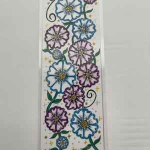 Könnte beinhalten: Ein rechteckiges Lesezeichen mit einer Blumenillustration aus blauen und violetten Blüten an grünen Stielen. Das Design enthält kleine goldene Sternakzente und gelbe Punkte auf weißem Hintergrund, alles in einer transparenten Schutzfolie.