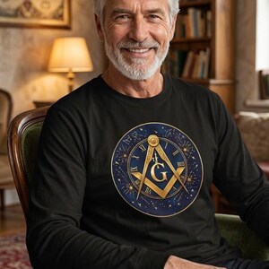 May include: A black long-sleeve shirt featuring a circular graphic on the chest. The design displays a gold Masonic square and compass symbol with the letter G, set against a dark blue background with Roman numerals and celestial star patterns.