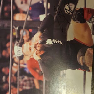 May include: A glossy photograph of a professional wrestler in a black long-sleeve shirt with a white fish skeleton graphic. The wrestler is kneeling in a ring with black ropes, wearing black knee pads with red accents and has a black signature on the image.