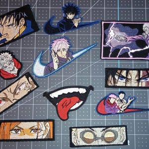 May include: A collection of embroidered fabric patches featuring various anime-style character illustrations, including close-ups of eyes, a mouth with a tongue, and characters integrated with swoosh shapes, all arranged on a gray grid mat.