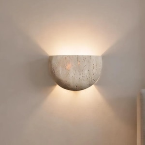May include: A semi-circular wall sconce with a textured stone finish emits a warm glow against a neutral wall. Below, a floating wooden nightstand holds a ceramic mug and a book. The logo for Midnight Beam Co is visible in the upper left corner.