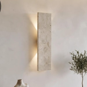 Puede incluir: Un aplique de pared rectangular con acabado de hormigón beige moteado montado en la pared, que emite luz desde los bordes superior e inferior. La lámpara se sitúa sobre un mueble flotante de madera clara con dos cajones y superficie lisa.