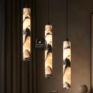 May include: Three cylindrical pendant lights with a marble-like pattern hang above a dining table. The lights have a black and white design and are suspended from the ceiling. A dark bowl, glasses, and a vase are on the table.