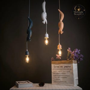 May include: Three squirrel-shaped pendant lights in navy, white, and peach. Each light has a decorative cap and a vintage-style bulb. The lights hang from white cords against a dark background. A paper bag with French text and purple flowers are in the foreground.